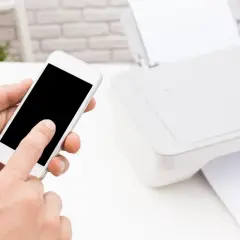 Dłoń trzyma smartfon, inicjując drukowanie z telefonu przez wifi. Obok drukarka z papierem.
