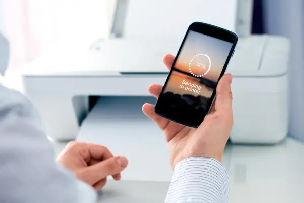 Dłoń trzyma smartfon z napisem "Sending to printer", pokazując jak drukować z telefonu. W tle biała drukarka.
