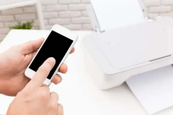 Dłoń trzyma smartfon, inicjując drukowanie z telefonu przez wifi. Obok drukarka z papierem.